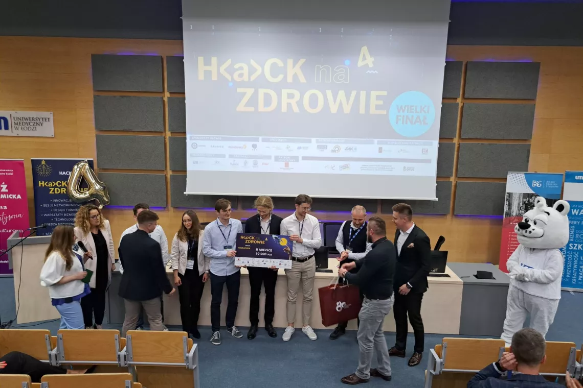 Osoby stoją przed ekranem z napisem Hack na zdrowie. Jedna z nich trzyma czek, inne osoby czekają na złożenie gratulacji