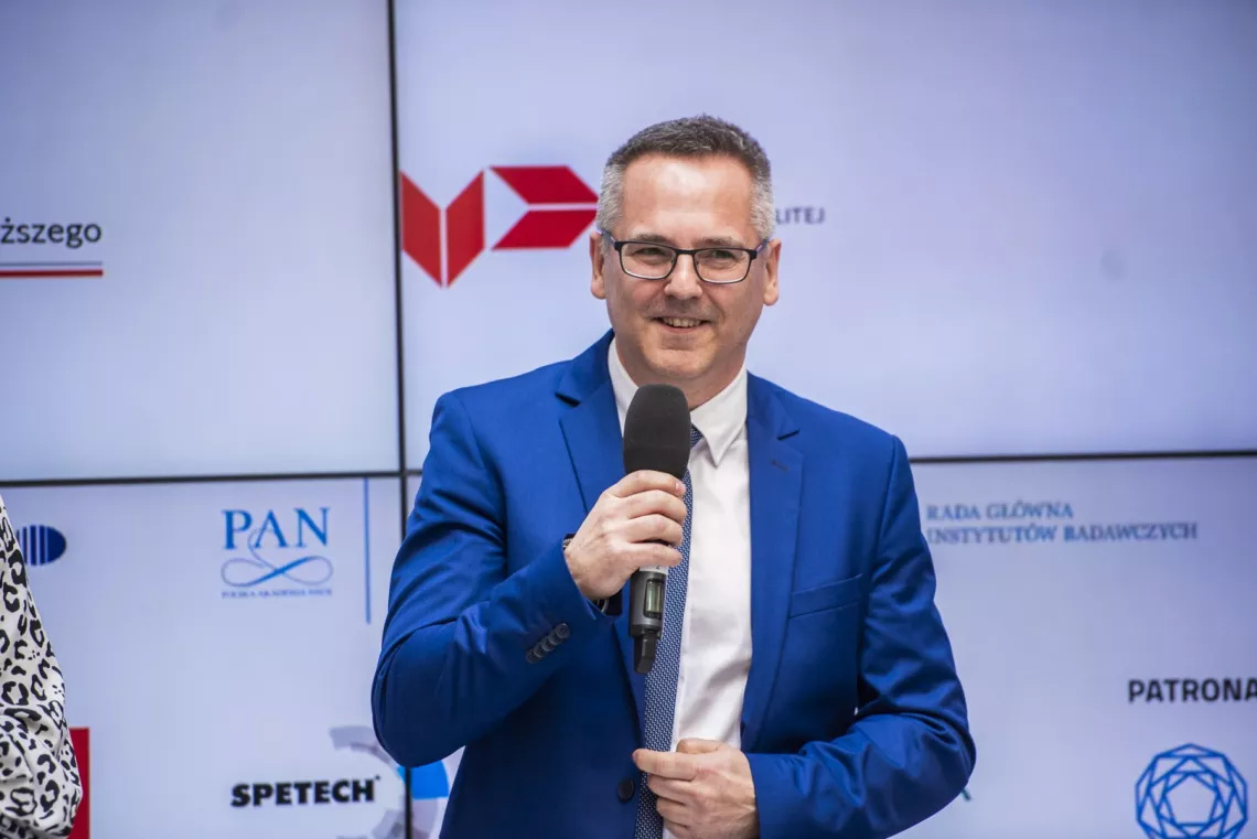 Osoba w niebieskim garniturze trzymająca mikrofon. W tle na ekranie znajdują się liczne loga.