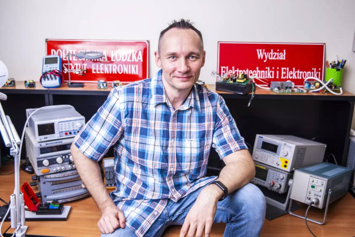 Osobę siedzącą przed biurkiem z różnym sprzętem elektronicznym i urządzeniami. Za osobą znajdują się czerwone tablice, po lewej stronie z napisem POLITECHNIKA ŁÓDZKA WYDZIAŁ ELEKTRONIKI, a znak po prawej  z napisem "Wydział Elektrotechniki i Elektroniki."