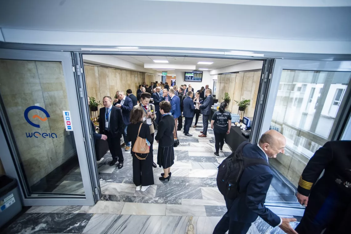 Wiele osób stoi w korytarzu i prowadzi rozmowy. Z przodu widoczne szklane drzwi z grafiką i napisem weeia, z prawej strony okna.