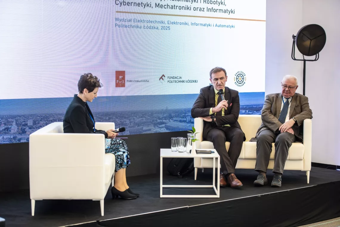 Trzy osoby siedzą na kanapach przy małym stoliku, jedna z nich wypowiada się do mikrofonu