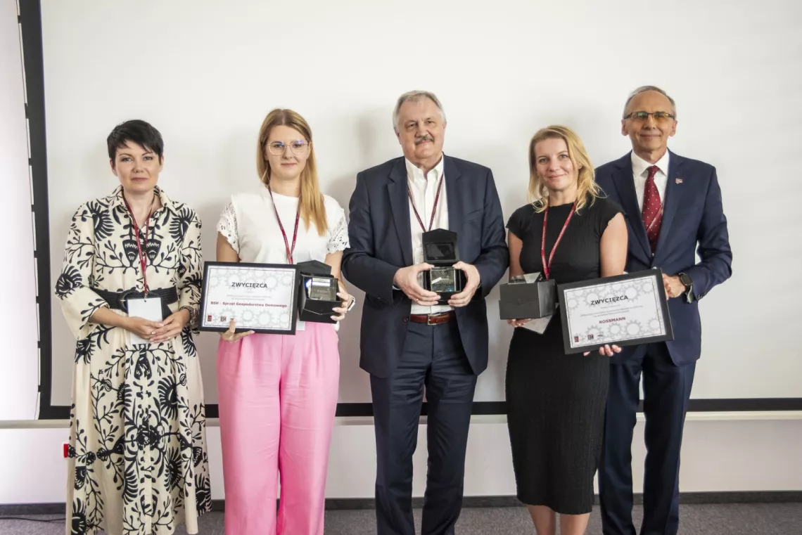 Trzy osoby stoją, trzymając nagrody i dyplomy, towarzyszą im dwie, które wręczały te nagrody