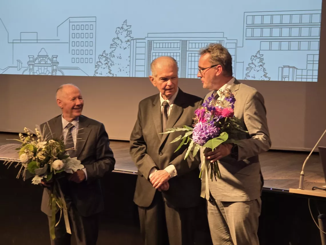 Trzej mężczyzni stojący na scenie, dwóch z nich trzyma w reku bukiet kwiatów