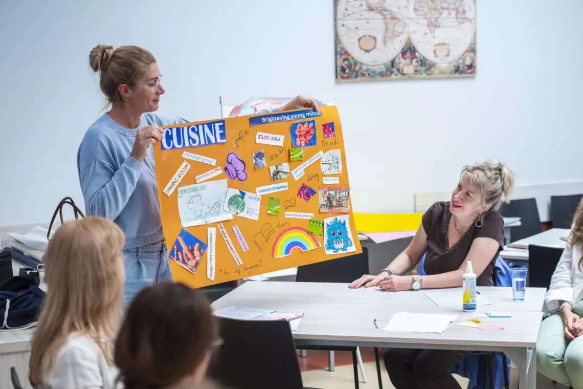 Kobieta trzyma w ręku pomarańczowy arkusz papieru, na którym naklejone są różne elementy. Stoi przy stole, przy którym siedzą inne osoby, a leżą na nim kartki, długopisy i rózne inne drobiazgi potrzebne do zrobienia prezentowanej pracy