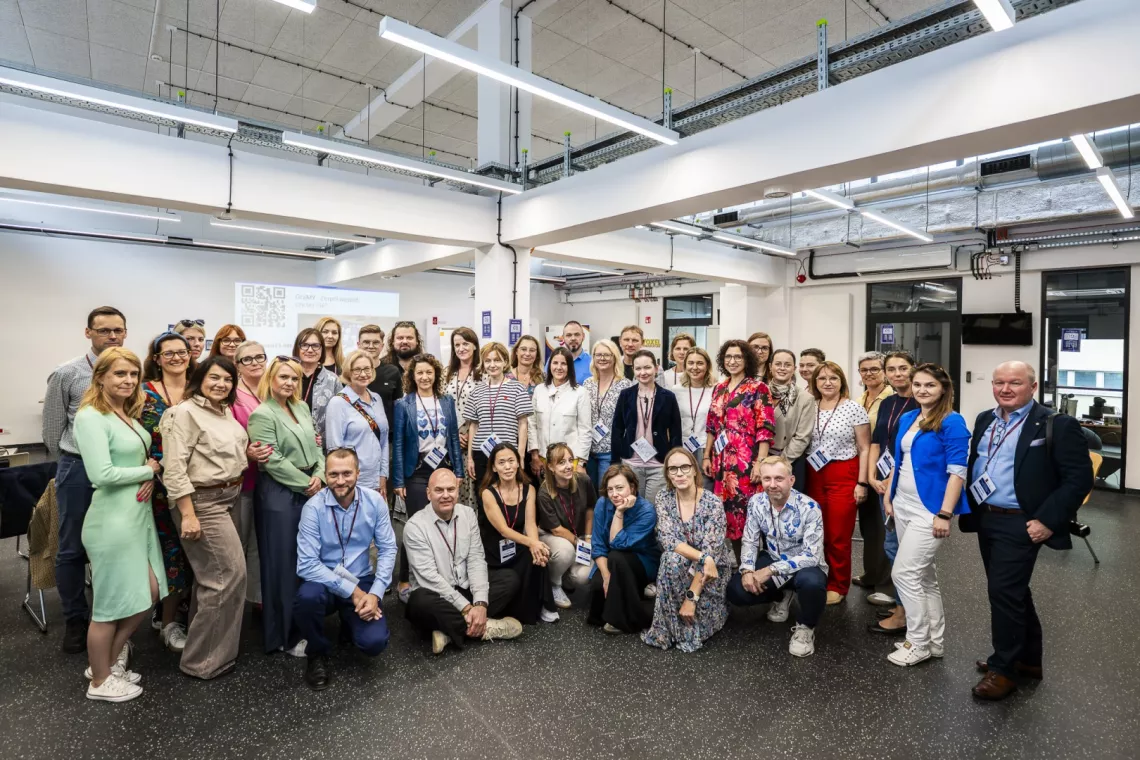 Do zdjęcia pozują uczestnicy konferecji stojący w otwartej przestrzeni, wnętrze ma charakter postindustrialny