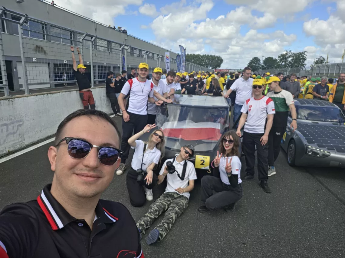 grupa młodych osób stoi wokół samochodu, trzy przed nim siedzą. Napierwszym planie osob robiaca selfie