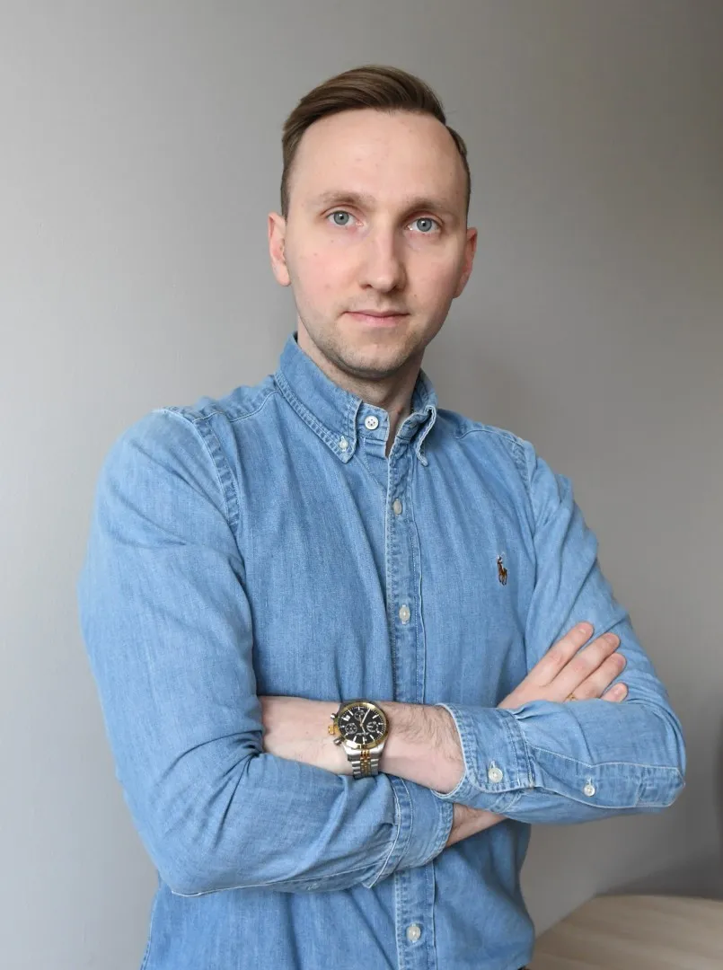 Młody mężczyzna w teksasowej koszuli, trzyma ręce skrzyżowane na ramionach, w tle szara ściana