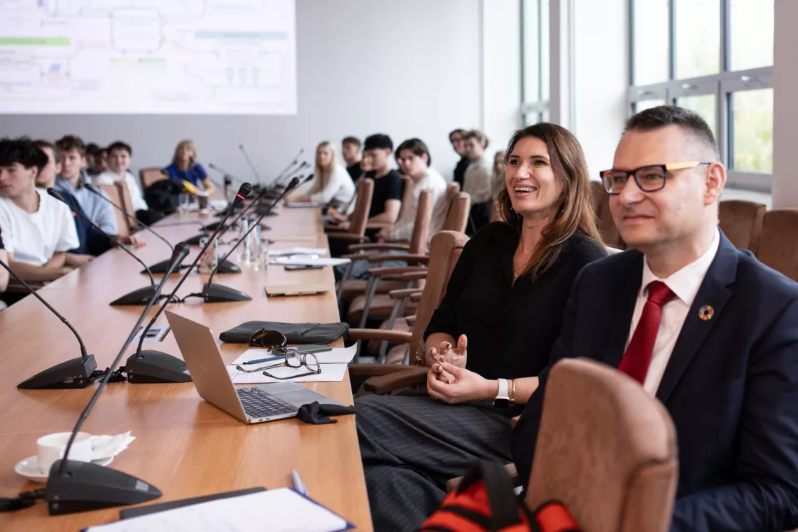 Grono osób siedzi przy długim stole w sali konferencyjnej, najbliżej widać uśmiechnięta kobietę, pzred którą leży otwarty laptop
