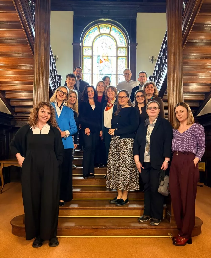 Kilkanascie osób stoi na schodach zabytkowego holu, z witrażem w tle