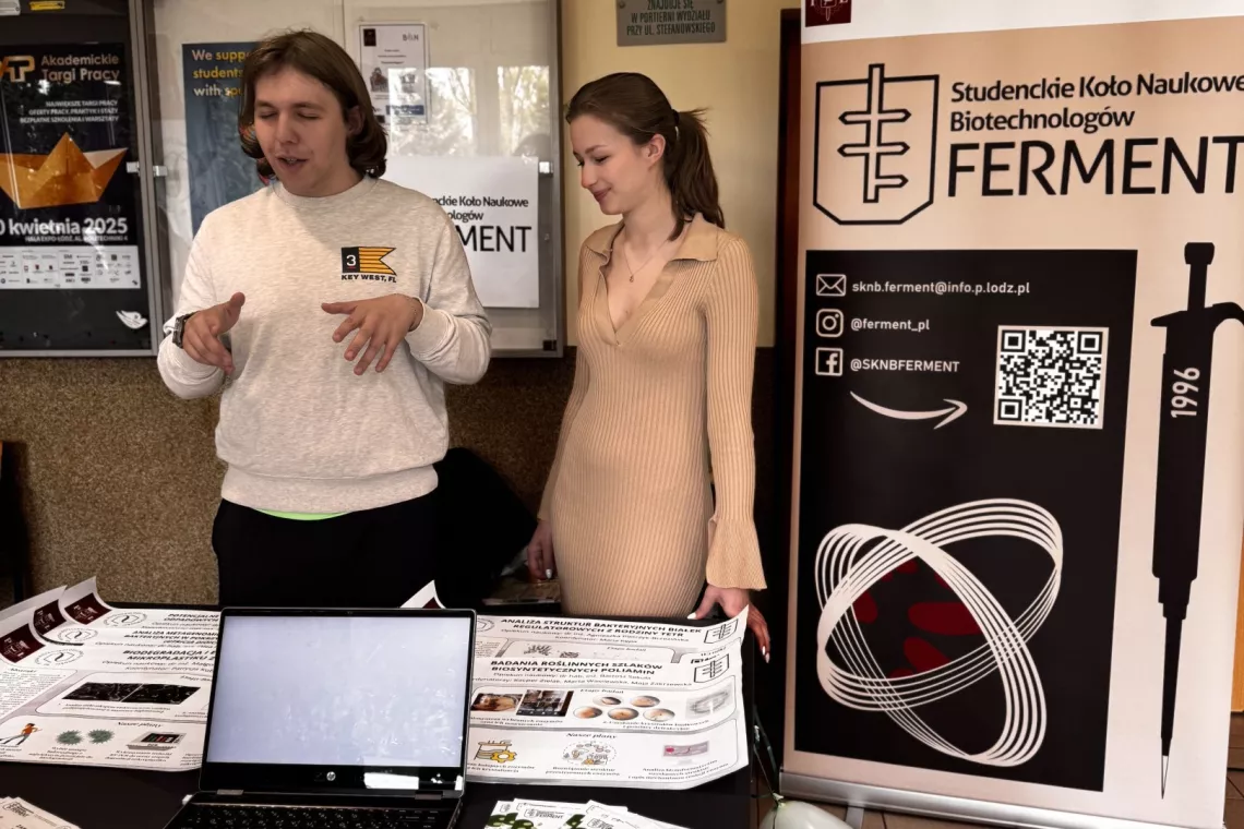 Dwie młode osoby przy stoisku, jedna z nich coś opowiada