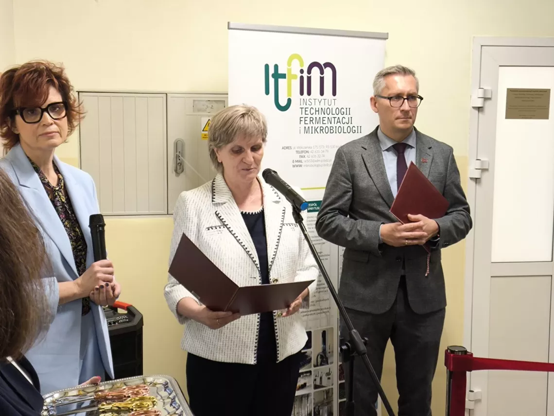 Dzwi kobiety i mężczyzna stoją przed wejścim do laboratorium, jedna z kobiet przemawia do mikrofonu