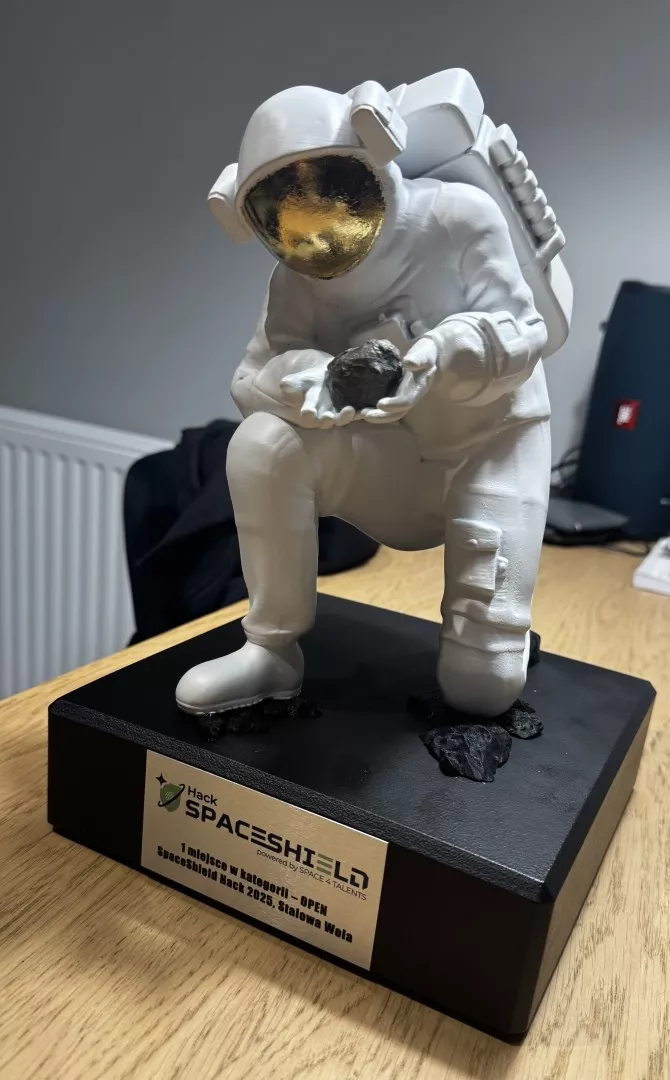 Statuetka kosmonauty w kafandrze kosmicznym, który klęczy, trzymając w dłoniach kawałek skały lub meteorytu. Statuetka umieszczona jes na czarnej podstawce