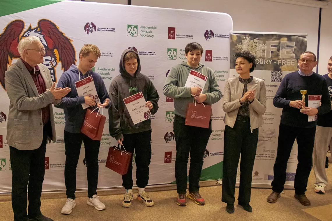 Grupa osób stojąca przed tłem z różnymi logotypami i tekstami. Cztery osoby trzymają certyfikaty i torby z upominkami, a jedna osoba trzyma trofeum. Dwie inne osoby stoją po bokach grupy, jedna klaszcze, a druga obserwuje. Na tle widoczne są logotypy Akademickiego Zrzeszenia Sportowego, HF Security-Free i innych organizacji