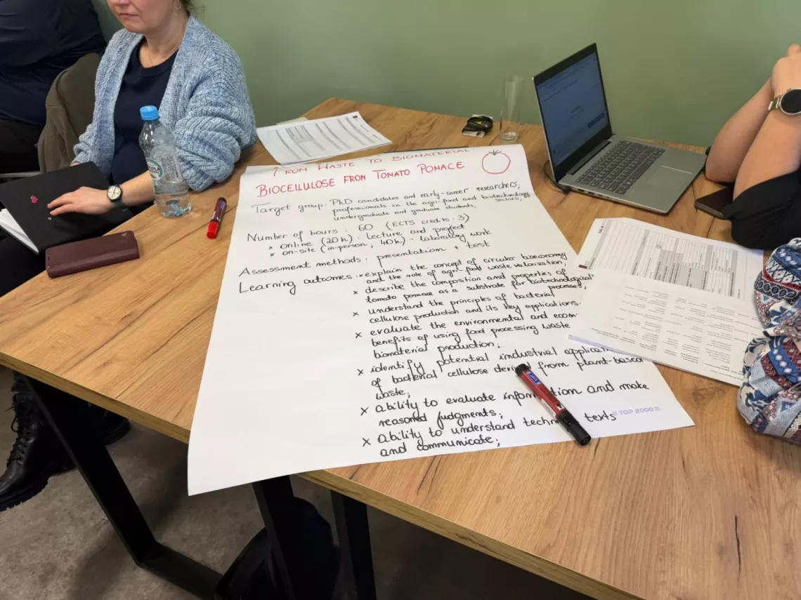 Tekst zapisany odrecznie na duzej kartce papieru leżącej na stole, przy którym widać kilka siedzących osób, na stole leży laptop