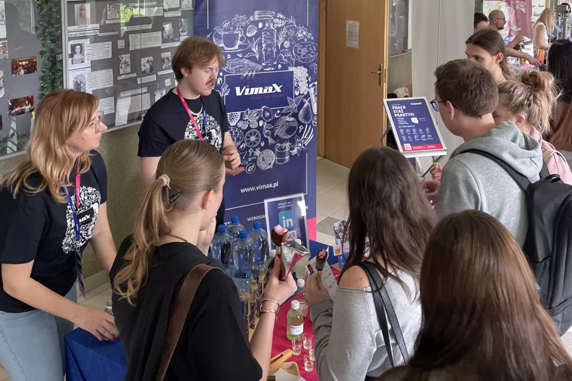 Grupa osób stoi i dyskutuje, w tle widać baner firmy