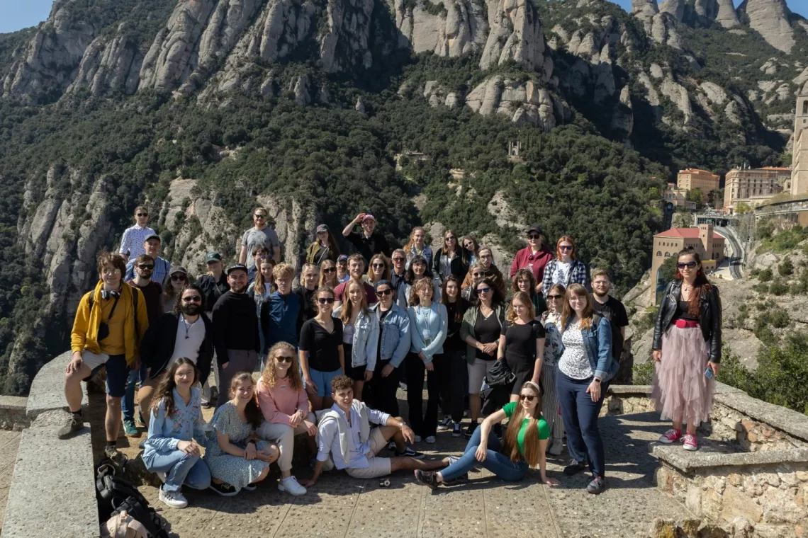 Duża grupa ludzi pozuje do zdjęcia na świeżym powietrzu, na tle górskiego krajobrazu. Grupa stoi i siedzi na kamiennej platformie, a za nimi widać góry pokryte drzewami i skalnymi formacjami. Po prawej stronie zdjęcia widoczne są budynki w oddali.