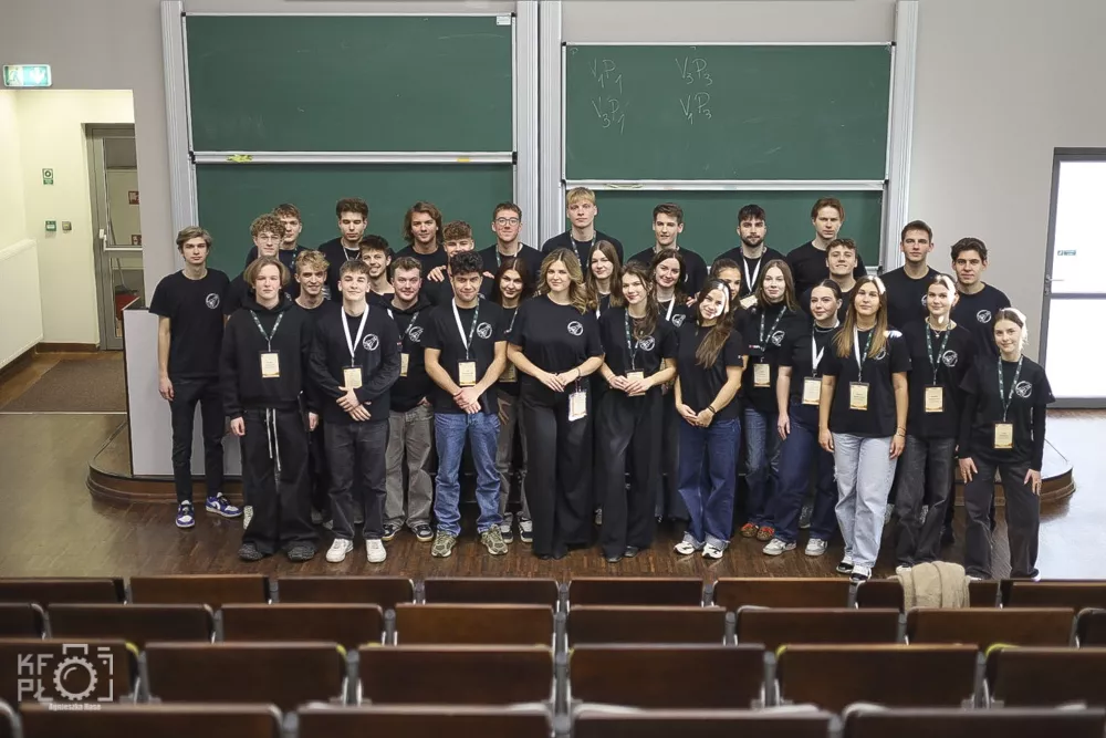 Grupa studentów i studentek w jednakowych koszulkach z identyfikatorami stoi na tle tablicy w audytorium