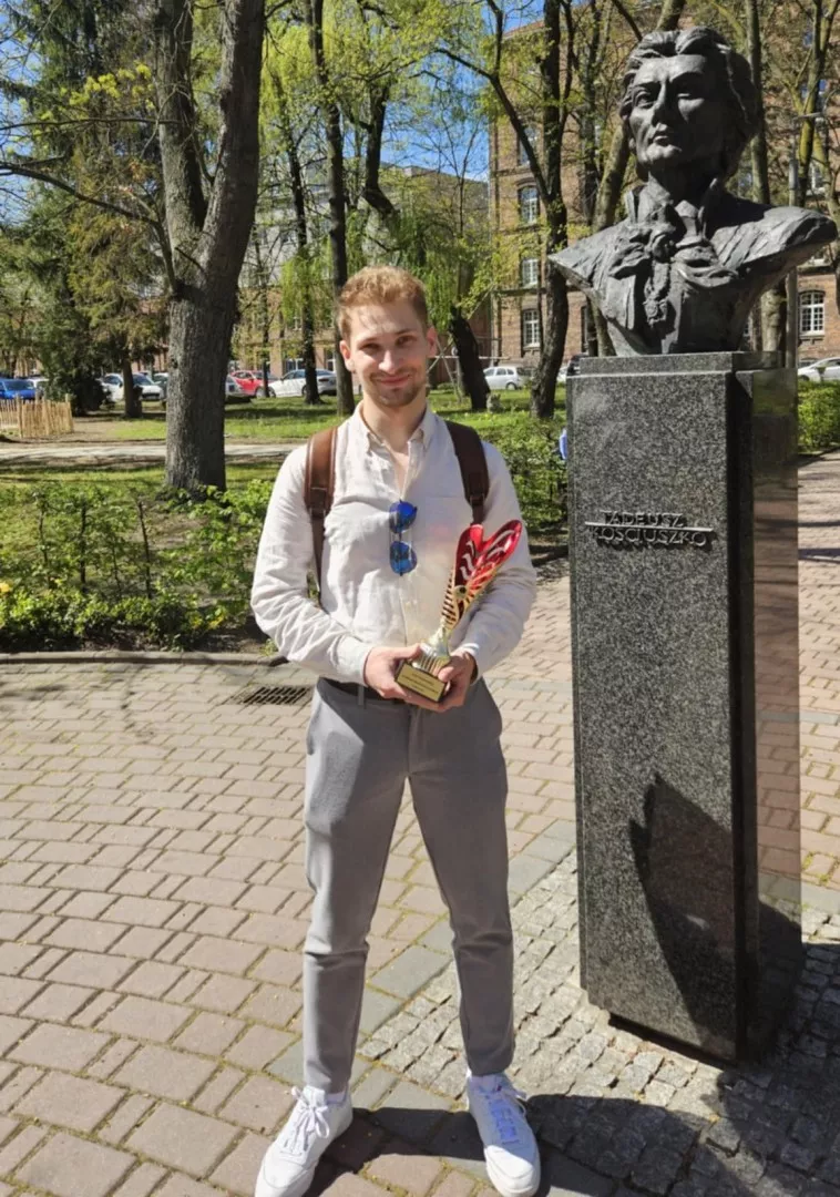 Młody mężczyzna w białej koszuli i szarych spodniach trzyma w reku puchar, stojąc przy pomniku z popiersiem Kościuszki