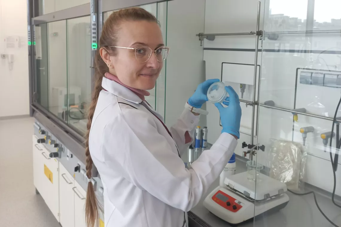 Kobieta w białym fartuchu trzyma w rękach próbke, stojąc pzry stanowisku laboratoryjnym