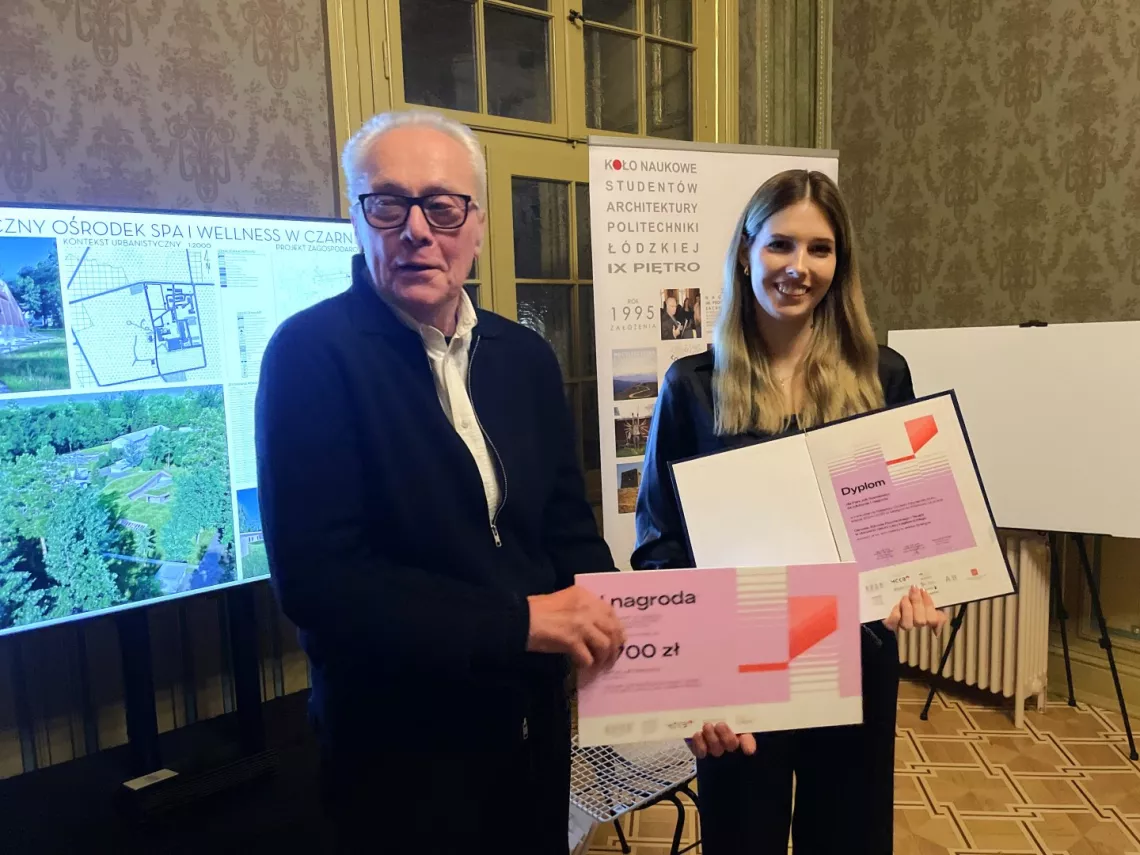 Młoda kobieta trzyma dyplom w ręku, obok mężczyzna z symbolicznym czekiem. Z tyłu praca architektorniczna na sztaludze i baner konkursu