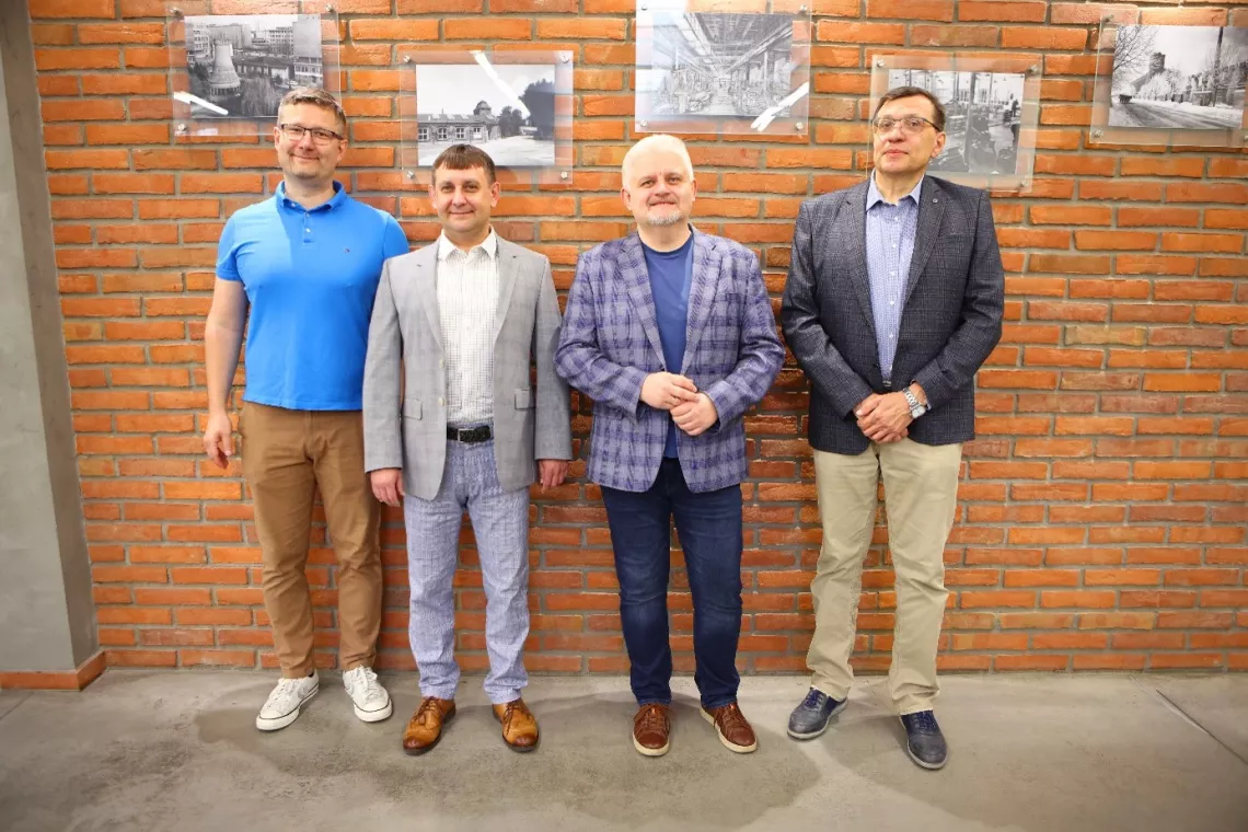 Czterech mężczyzn stoi na tle ceglanej ściany, na której wiszą czarno-białe fotografie