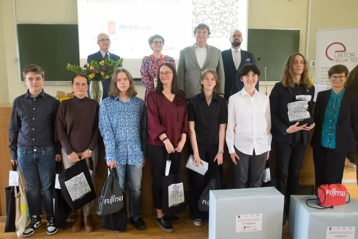 Kilkoro młodych osób stoi z torbami materiałowymi, zapewne zawierającymi magrody. Z tyłu, za prezydium stoi czworo elegancko ubramych  dorosłych osób.