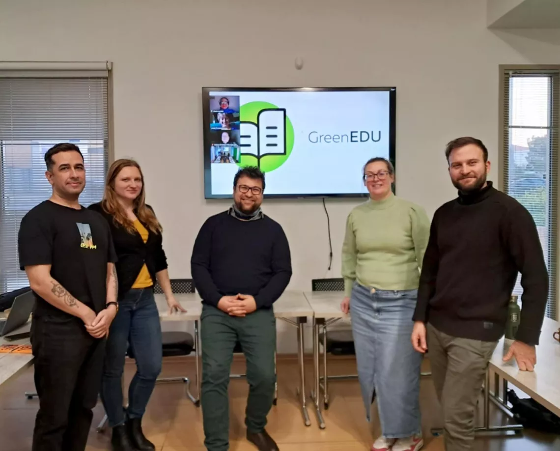 Pięć osób stoi w pokoju z dużymi oknami. Na ekranie telewizora za nimi widnieje logo i nazwa 'GreenEDU'