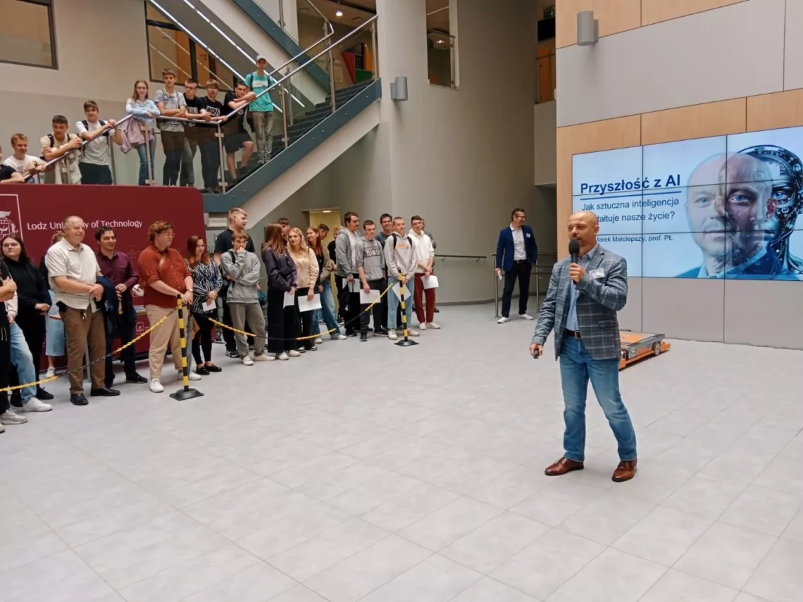 Wykładowca stoi w otoczeniu słuchających, z tyłu na ekranie tytuł wykładu Przyszłość z AI