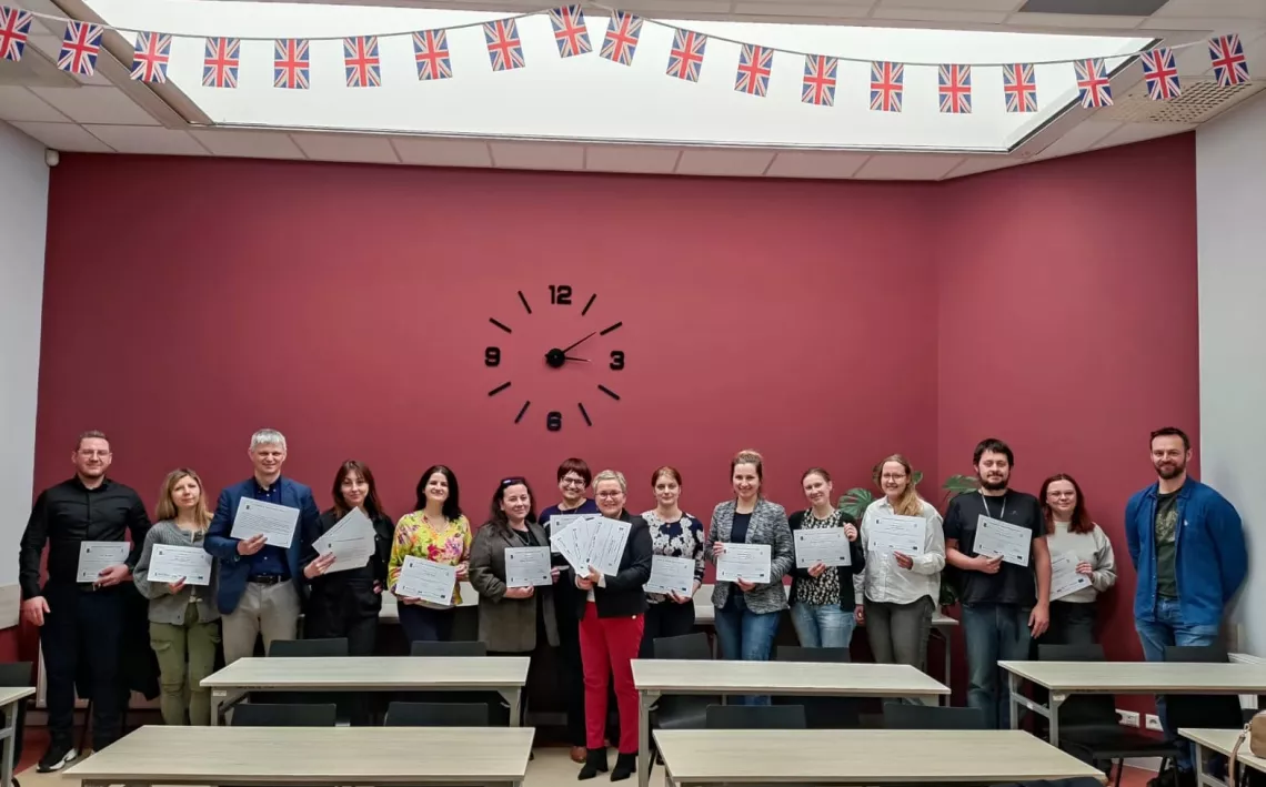 Kilkanaście osób z dokumentem w ręku stoi na tle bordowej ściany z zegarem