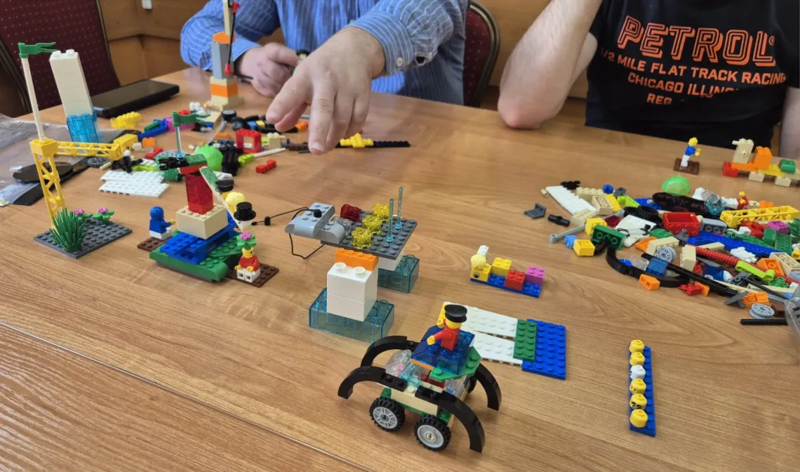Różne konstrukcje z klocków Lego leżą na stole, pzry którym są także, widoczne w części, dwie osoby