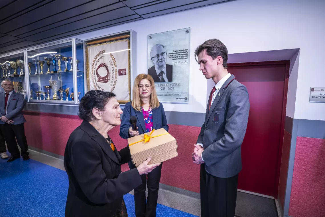 Kobieta  w czarnej marynarce, ze spiętymi czarnymi włosami trzyma w ręku pudełko przewiązane kokardą i wręcza je wysokiemu uczniowi w szarym szkolnym mundurku, obseruje ten moment stojąca obok kobieta z jasnymi długimi włosami, w granatowej marynarce i kolorowej koszuli.