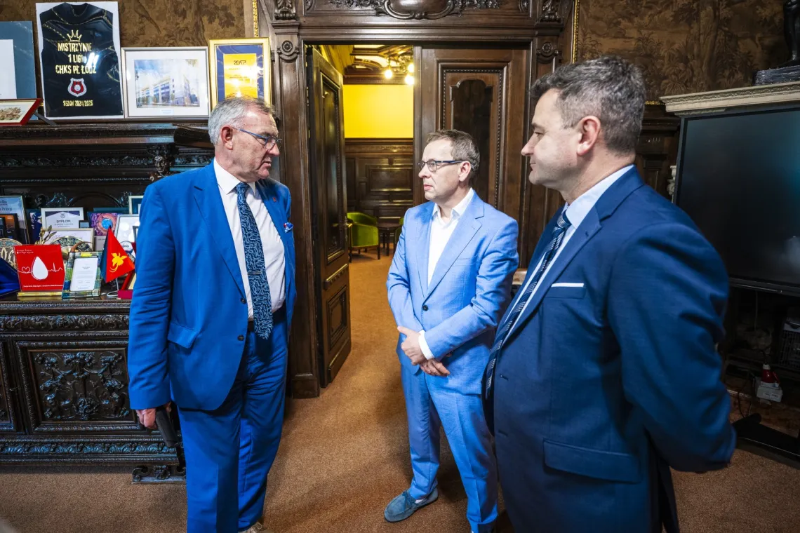 Trzech elegancko ubranych mężczyzn prowadzi rozmowę w gabinecie z historycznym wystrojem