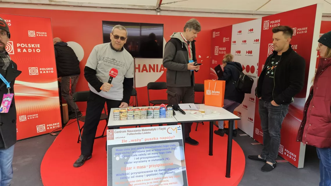 Osoba z mikrofonrm trzyma mikrofon, stojąc przy stole na którym stoją puszki. Całość umieszczona jest pod namiotem wystawowym graficznie oznaczonym Polskie Radio
