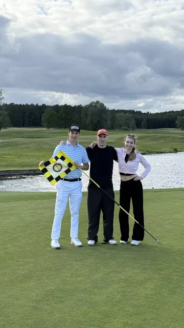 Trzy osoby stoją na polu golfowym, jedna z nich trzyma chorongiewkę, z tyłu widoczny dołek z wodą 
