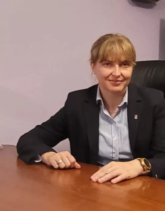 Koebita siedzi przy stole, trzymając ręce na blacie. Jest ubrana w marynarkę i koszulę.