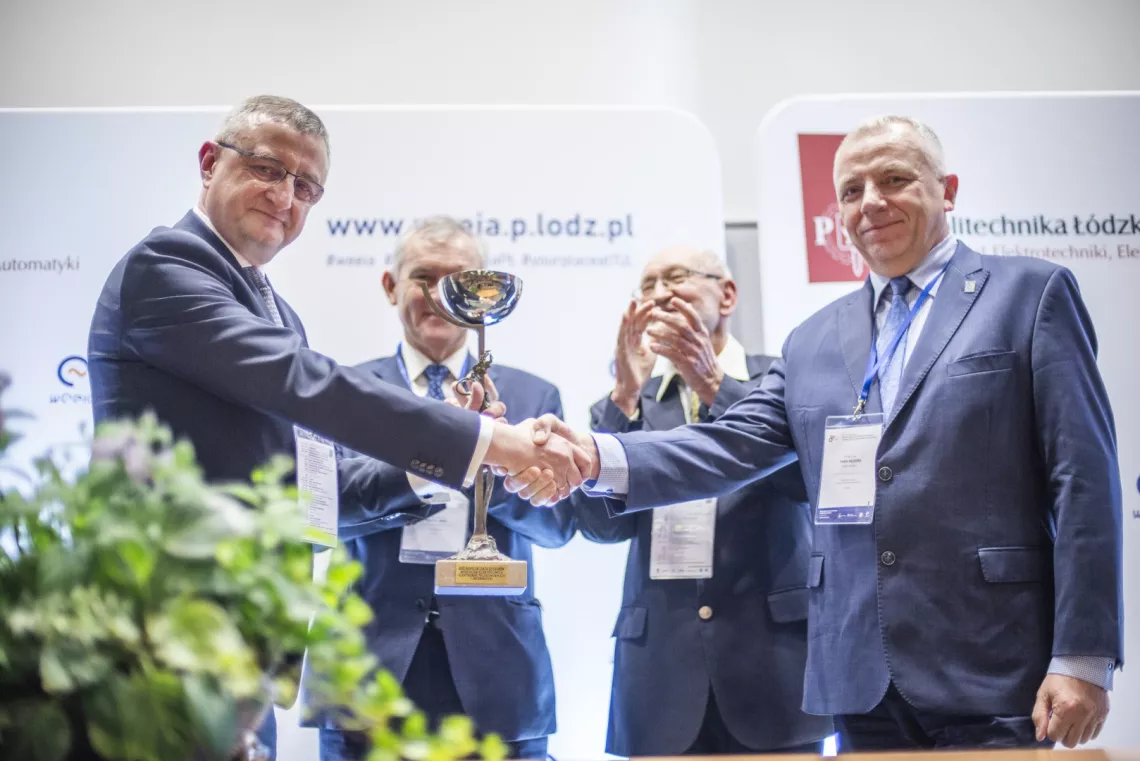 Przekazanie pucharu przez jedną osobę drugiej osobie. Z tyłu przyglądają się temu dwie inne osoby, jedna z nich klaszcze.