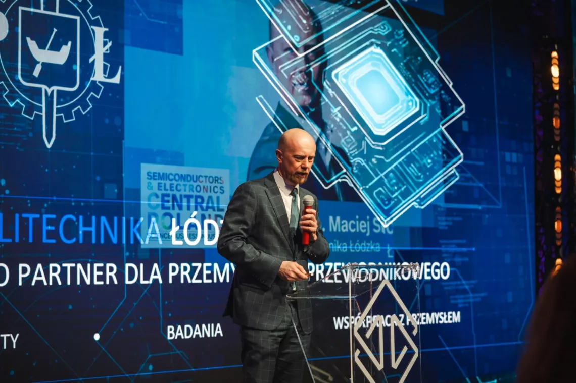 Przemawia mężczyzna z mikrofonm w ręku. Z tyłu duży ekran wyświetlający logo PŁ, grafike czipa oraz tekst Politechnika Łódzka partner dla przemysłu pólprzewodnikowego