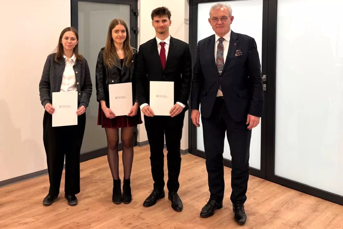 Troje studentów studentów stoi, trzymając w ręku dyplomy, towarzyszy im elegancko ubrany mężczyzna w garniturze