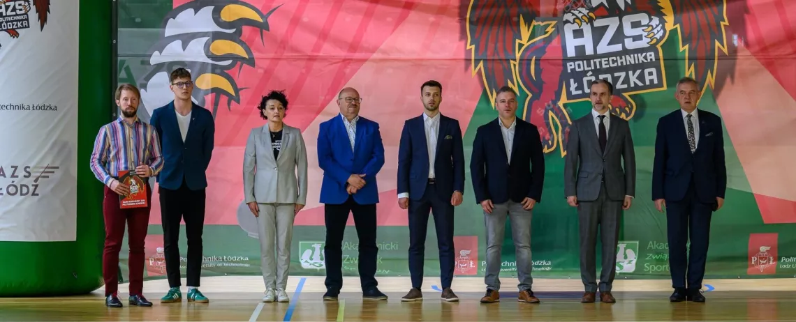 Osmioro elegancko ubranych osób stoi, zachowując poważną pstawę. W tle baner z napisem AZS Politechnika Łódzka.
