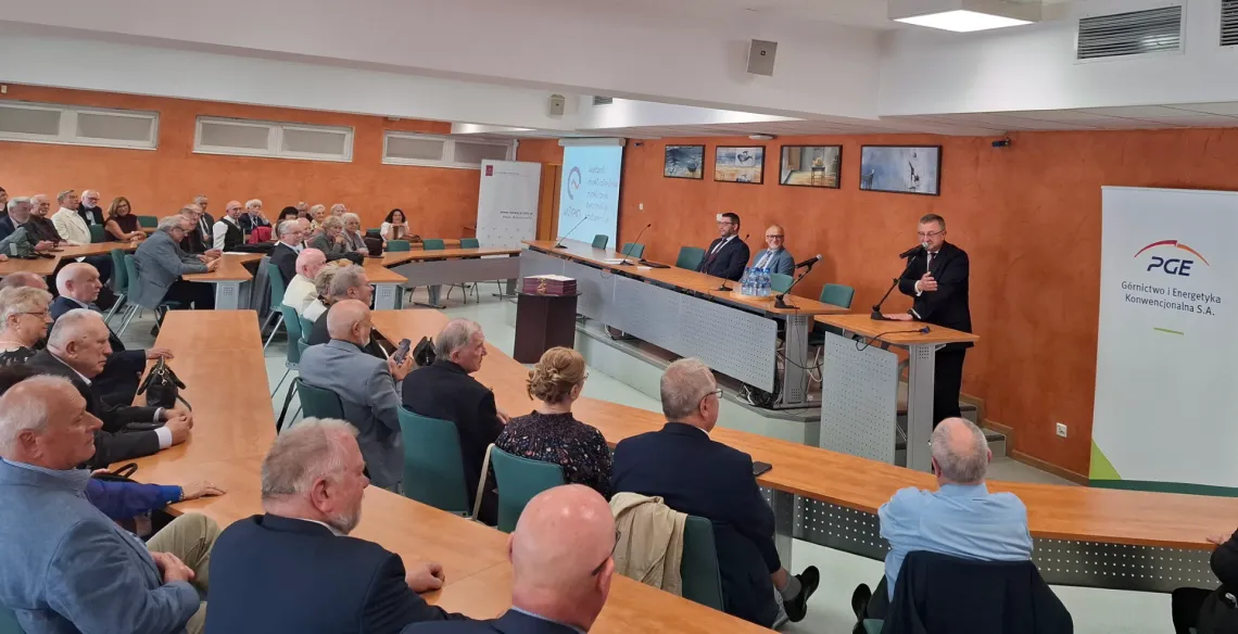 Sala konferencyjna wypełniona uczestnikami, za stołem prezydialnym siedzą dwaj mężczyżni, przy mównicy stoi mężczyzna w ciemnym garniturze