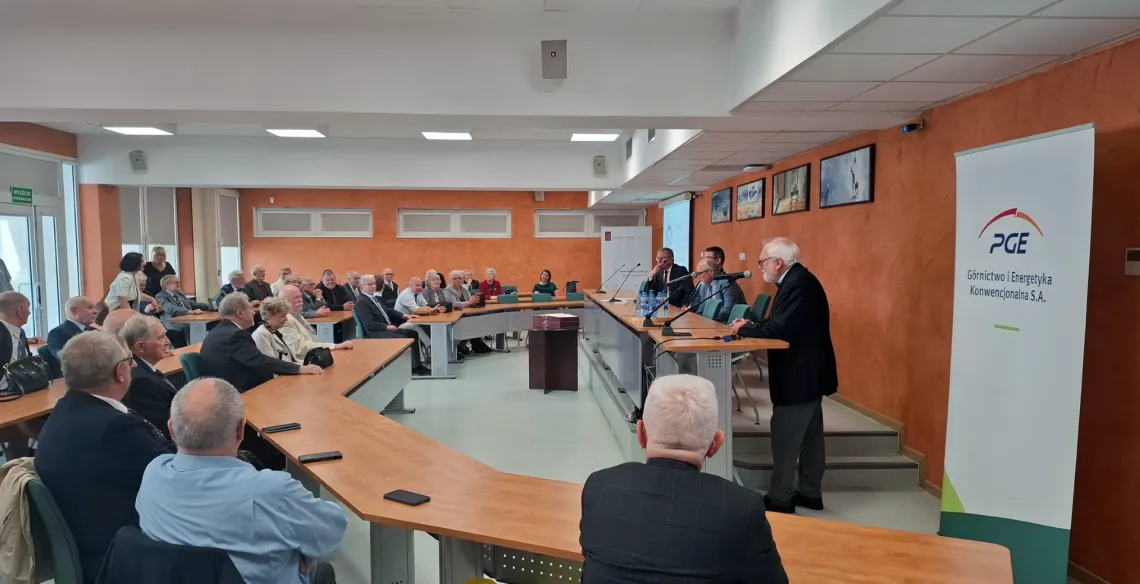 Sala konferencyjna wypełniona uczestnikami, za stołem prezydialnym siedzą trzej mężczyżni, przy mównicy stoi mężczyzna z siwymi włosami, obok rollup z napisem PGE Górnictwo i Energetyka Konwencjonalna S.A. 