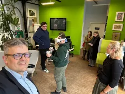 Kilka osób zebranych w pokoju z zielonymi ścianami, obrazkami na scianach i roślinami obserwuje mężczyznę w okularach do VR niosącego kubki z napojami