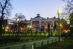 Zabytkowy budynek w otoczeniu zielenie, o zmroku, świecą się uliczne latarnie i okna w budynku