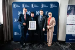 Dwie kobiety - jedna z nich trzyma dyplom - i dwóch mężczyzn stoi na tle ścianki z napisem Polska Komisja Akredytacyjna