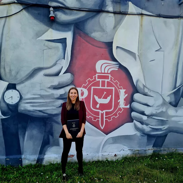 Uśmiechęta młoda kobieta stoi na tle muralu z logo PŁ