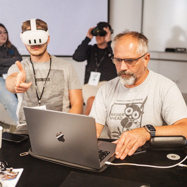 Mężczyzna pracuje przy laptopie, obok student z goglami VR