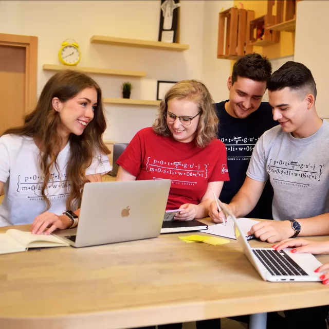 Grupa studentów siedzi pzry stole, na którym stoją laptopy i rozłożone kartki.  Prowadzą ze soba ozywioną dyskusję.