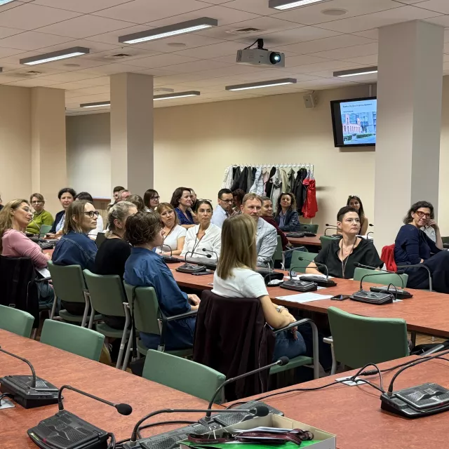 Uczestnicy konferencji siedzą w sali seminaryjnej, na stołach leżą urządzenia z mikrofonami z tyłu stoi wieszak na ubrania, na ścianie wisi monitor