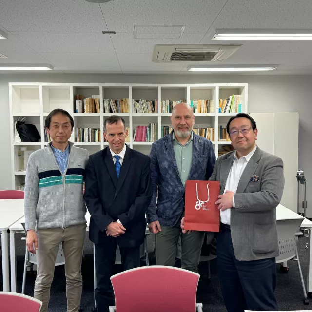 czterech mężczyzn stojących w biurze przed półkami z książkami. Jeden z nich trzyma czerwoną torbę z białym symbolem. W tle znajdują się stoły, krzesła i tablica suchościeralna.