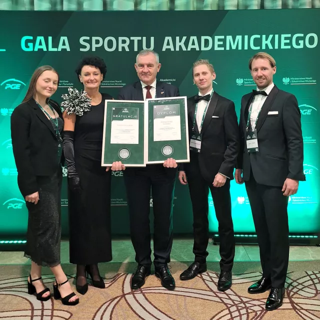 Pięć osób eleganko ubranych, jedna z nich, stojaca w środku trzyma w ręku dwa dyplomy. Z tyłu ścianka z napisem Gala Sportu Akademickiego